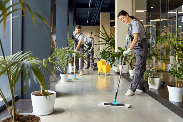 Nettoyage de bureaux : trois personnes en salopette grise lavent et aspirent le sol d'un couloir vitré bordé de plantes